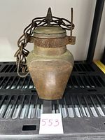 Front view of antique hand hammered copper camel canteen placed on black shelf with chain attached and visible rust on the locking ring at top.