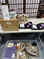 Overall view of table with wooden crate holding various craft supplies, two black cases with pink hot melt glue guns, wooden decorative houses, and a plastic bin full of diverse craft materials.