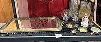 Photo of large vintage wall mirror with ornate gold floral frame, and assortment of smaller brass frames, glass candle holders, and porcelain figurine on black surface.