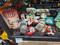 Photo showing entire lot including Santa themed metal latched cookie tin, small pre-lit Christmas tree with decorations, holiday candle in glass holder, ceramic Santa footed platter, and assorted Christmas ornaments including figurines and baubles.
