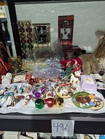 Wide view of assorted Christmas ornaments and decorations laid on table, showing colorful glass ball ornaments, figurines, plates, and packaged nutcracker doll.