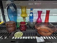 Overall display of assorted colored glassware on metal shelving, showing a variety of vases, candle holders, plates, and containers in multiple glass colors.