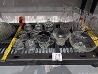 Wide view of all glassware items arranged on a black rack, showing pitchers, covered butter cups, sugar and creamer set, and footed glass bowls.