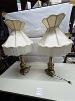 Pair of two brass lamps with cream fabric tassel lampshades placed on a white table with a lot number card in front