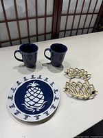Top-down view of brass pineapple trivet, blue and white Curzon plate, and two blue Royal Norfolk mugs on white surface