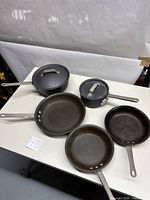 Five pieces of cookware including two saucepans with lids and three frying pans on a white table.