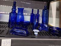 Full lot overview showing all blue glassware pieces including bottles, vases, bowls and covered dish arranged on black shelving.