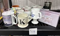 Photo of assorted coffee mugs including souvenir mugs and the cow creamer in its box on a black shelf.