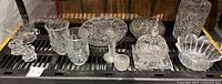 Collection of assorted crystal and pressed glass items including candle holders, footed dish with lid, vases, and bowls arranged on a shelf.