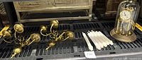 Photo showing brass clock under glass dome, two brass wall candle sconces, six brass candlesticks, and white taper candles.