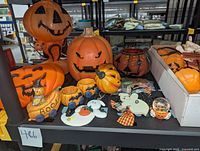 Wide shot showing a variety of Halloween ceramic pumpkins, including large pumpkins with carved faces, a ceramic pumpkin tealight train, and some wooden flat Halloween ghost and pumpkin decorations on a black shelf.