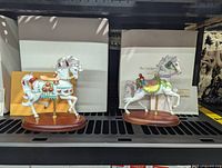 Two Lenox porcelain carousel horse figurines on wooden bases, each accompanied by original boxes and COAs. The horses are decorated in patriotic and holiday themes.