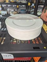 Stack of 11 white Lenox dinner plates with embossed rim design on metal shelf.