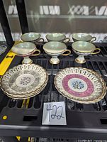 Front view of four porcelain pedestal cups with mother-of-pearl finish and four matching pierced saucers with floral patterns and gold accents