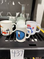 Front view of three novelty mugs featuring Alice in Wonderland, Raggedy Ann and Andy, and Avon 1969 Charlie Brown character designs, in front of milk glass pieces including hobnail jar and vase.