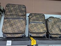 Three Oleg Cassini suitcases in brown plaid pattern lined up on a shelf showing front view with leather trim and logo.