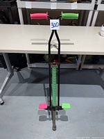 Front view of pogo stick showing neon pink and green handles and footrests, black frame, and coil spring.