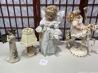 Blonde porcelain doll in white dress, wooden angel figurine, and ivory plastic angel lamp shown together