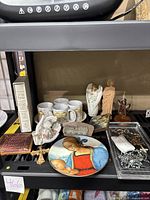 Shelf displaying the Precious Moments mugs, tray, Holy Bible, Last Supper plaque, angel figurines, Saint Jude statue, commemorative plate, and other assorted religious decor items.