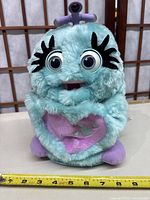 Front view of Trendmaster Aqua Wuv plush toy showing aqua blue plush with large eyes, black eyelashes, purple feet, ears, and heart emblem detail
