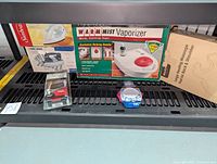 Photo showing the Robitussin Warm Mist Vaporizer box with details of warm soothing vapor and an exclusive helping handle, placed on a shelf with other items.