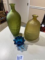 Photo showing the three glass pieces: tall green satin vase, yellow barrel can reproduction, and small blue artichoke flowerette vase on a white table.