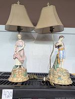 Full view of pair of vintage porcelain figure table lamps with beige shades, showing both lamps side by side.