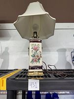 Full view of vintage mid century table lamp with floral painted ceramic base, gold trim, and white fabric shade, sitting on a black surface.