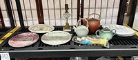 Wide view of lot showing various tea pots, plates, lamp, doll and small items on a black shelf
