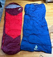 Two sleeping bags laid flat on wooden floor, one red and maroon mummy style, one blue rectangular style with plaid interior visible.