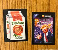 Two front-facing stickers laid flat on wood surface, showing parody art of Donald Trump as product and video game.