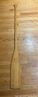 Full-length photo of the wooden paddle lying on a wooden floor showing the side with a red and white circular logo near the blade.