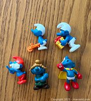 Top down image of 5 Smurf figurines on wood floor showing front profile with close detail on colors and accessories