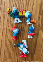 Top view of five vintage Smurf toys showing details including gift box, flowers, trumpet, wand, accordion, and telescope.