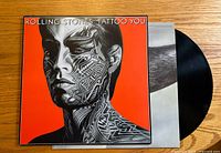 Front cover of the vinyl record album Tattoo You by The Rolling Stones, showing tattoo-style artwork on orange background and part of the vinyl record visible.