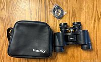 Black Tasco binoculars with central focus wheel, rubber grip, and attached neck strap. Binoculars placed next to a black Tasco zippered carrying case and separate plastic-wrapped neck strap.