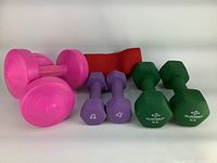 Pink, purple, and green dumbbells placed together with a red resistance band on a white background.