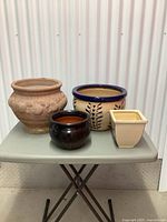 Photo showing four assorted vintage pots arranged on a gray folding table against a corrugated metal background.