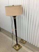Full view of the floor lamp showing beige fabric shade, dark wood body, brass accents on pole and base, standing on a concrete floor.