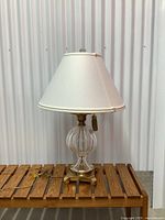 Front view of a vintage crystal table lamp with brass base, vertical ribbed crystal body, white fabric lampshade with beige trim, and pull chain with tassel. Lamp is on wooden slat surface against corrugated metal background.