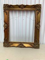 Front view of a large ornate gilded wood picture frame standing against a white corrugated metal wall.