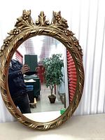 Front angle view of the full oval mirror showing the gold ornate floral frame and mirror surface reflecting a person and storage units.