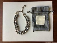 Double strand faux pearl necklace displayed next to branded soft pouch and certificate of authenticity on neutral background.