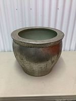 Front and side view of a large round metal pot with a wide rim and textured silver-tone exterior with pale green interior coating, placed against a white corrugated background.