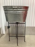 Front view of large galvanized steel bucket mounted on a black metal stand with four legs, placed in front of corrugated metal wall and metal diamond plate panel on floor edge.