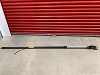 Full length of the electric trimmer with long black pole and black cutting head with orange grip and carrying strap shown on concrete floor.