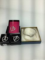 Photo showing three heart pendants with initial charms 'F', 'M', and one without initial, in individual boxes, and one silver-tone V-shaped bangle bracelet in a box with cushioning.
