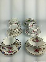 Six fine china teacups and saucers arranged in two rows, showing various floral patterns and gold trim.
