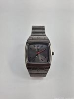 Front view of men's NIXON watch showing dark grey metallic square case with black dial, silver hour markers, and red second hand.