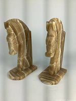 Pair of vintage hand carved horse head book ends made of solid stone with brown and beige banding patterns. Both standing on flat bases, one slightly loose with minor chips visible.
